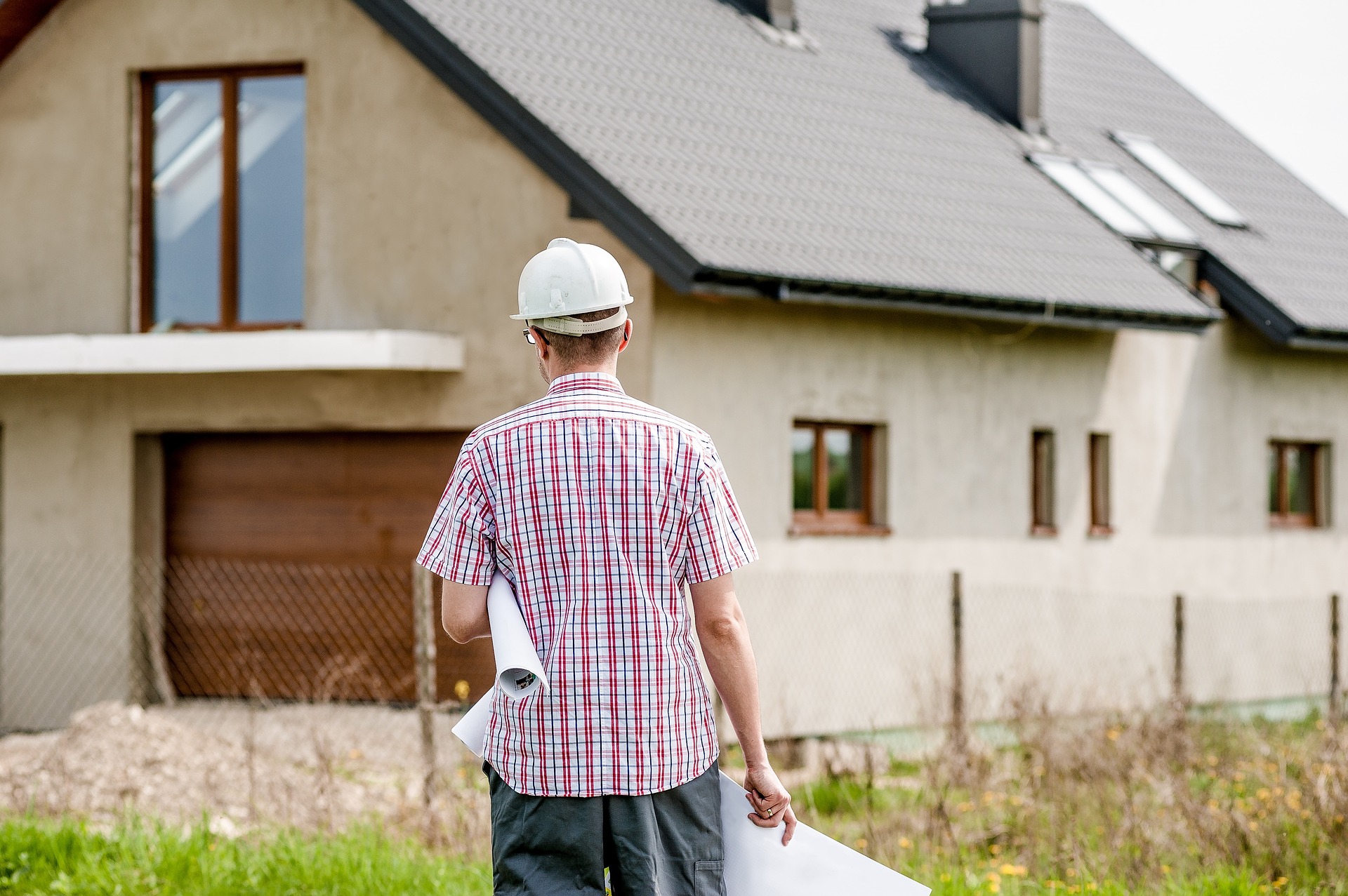 Photographie d'un homme de dos, portant des plans et un casque de chantier et faisant face à une maison